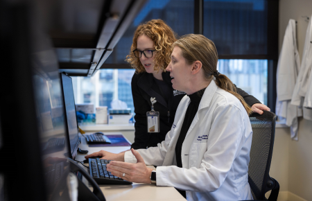 Two women, one sitting in an office chair looking at a computer and another woman leaning over her shoulder to look at the same computer screen.